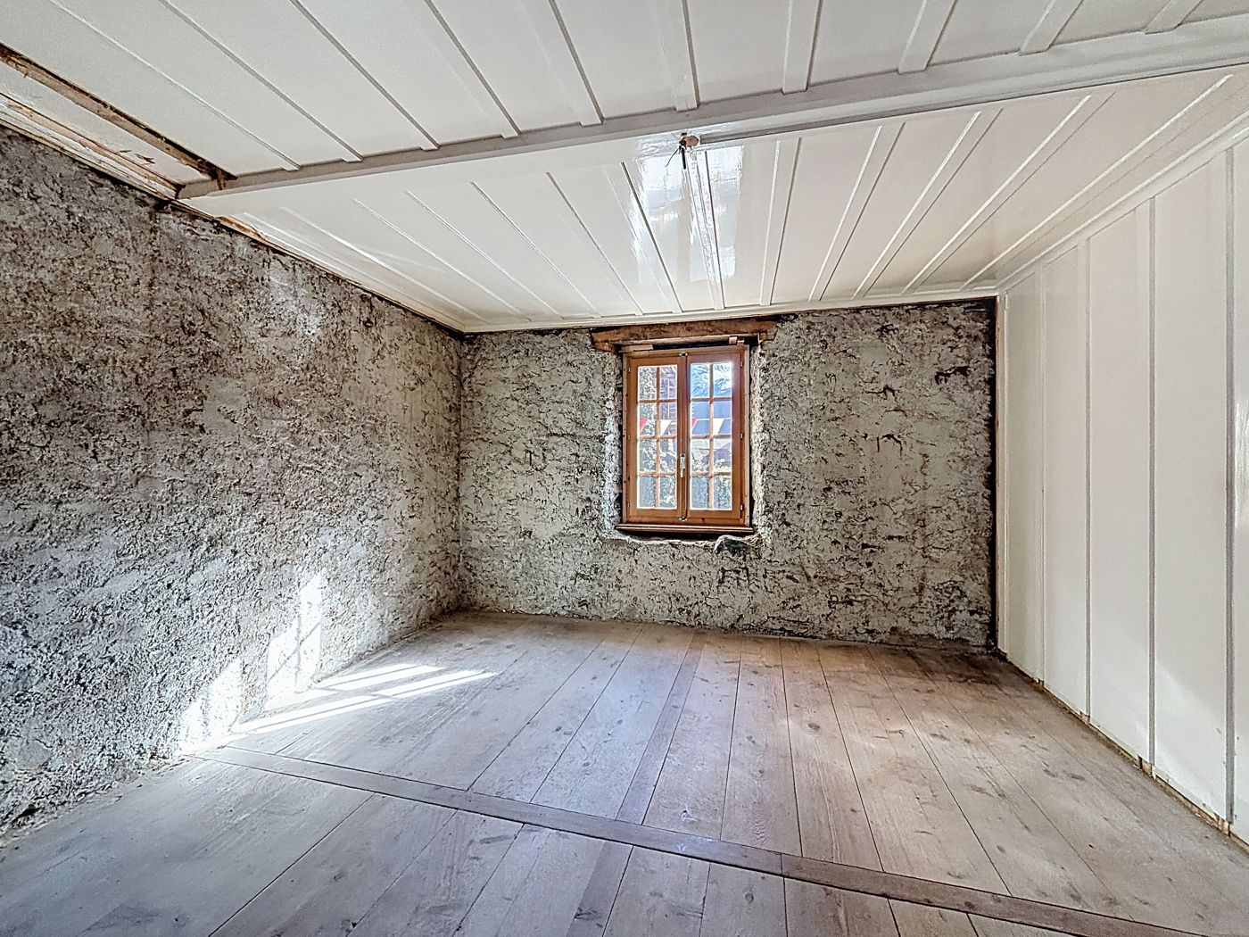 Pièce vide aux murs de pierre et parquet large, lambris blanc au plafond et sur la paroi droite, fenêtre au fond.