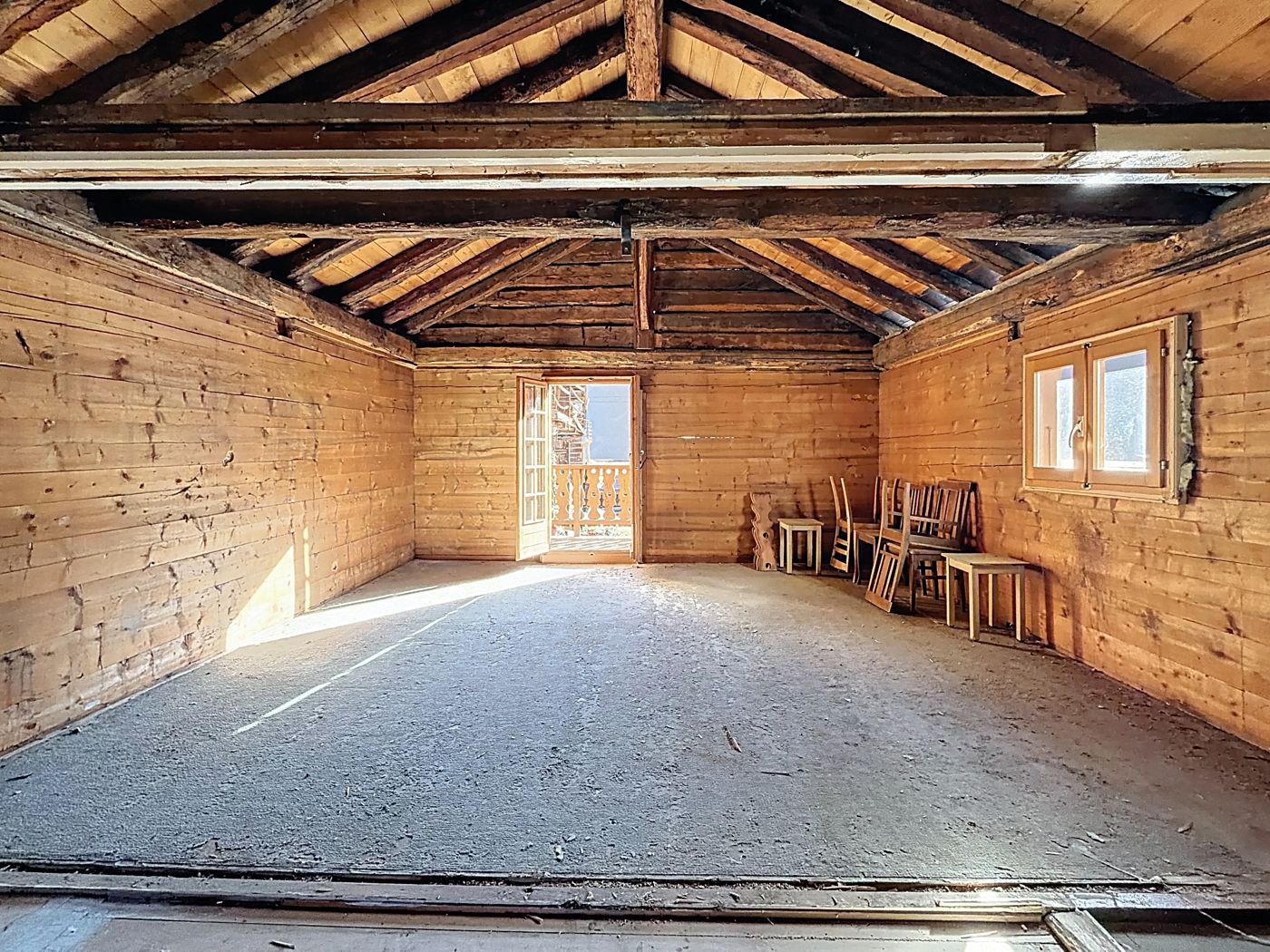 Le dernier niveau de l'ancienne grange : charpente apparente, madriers blonds, soleil entrant par la porte du balcon.