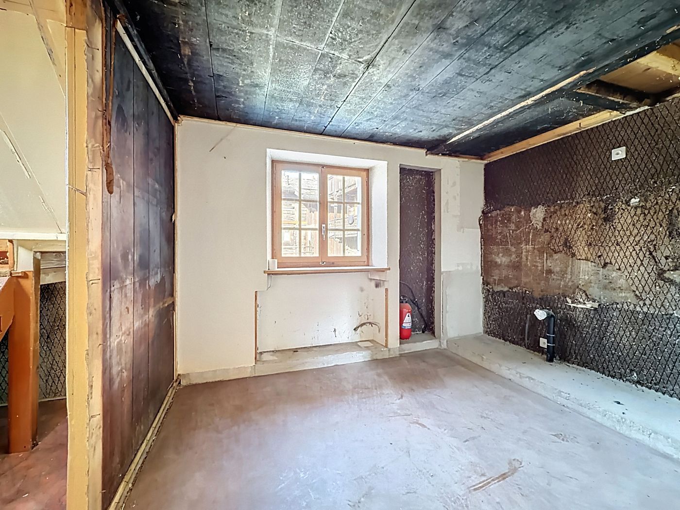 Pièce en travaux : fenêtre à petits carreaux, plafond en bois sombre, parois partiellement dégarnies.