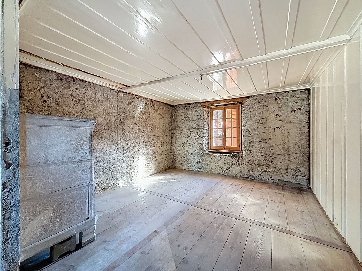 Pièce aux murs en pierre, fenêtre à petits carreaux, plafond lambrissé blanc, ancien buffet à gauche.