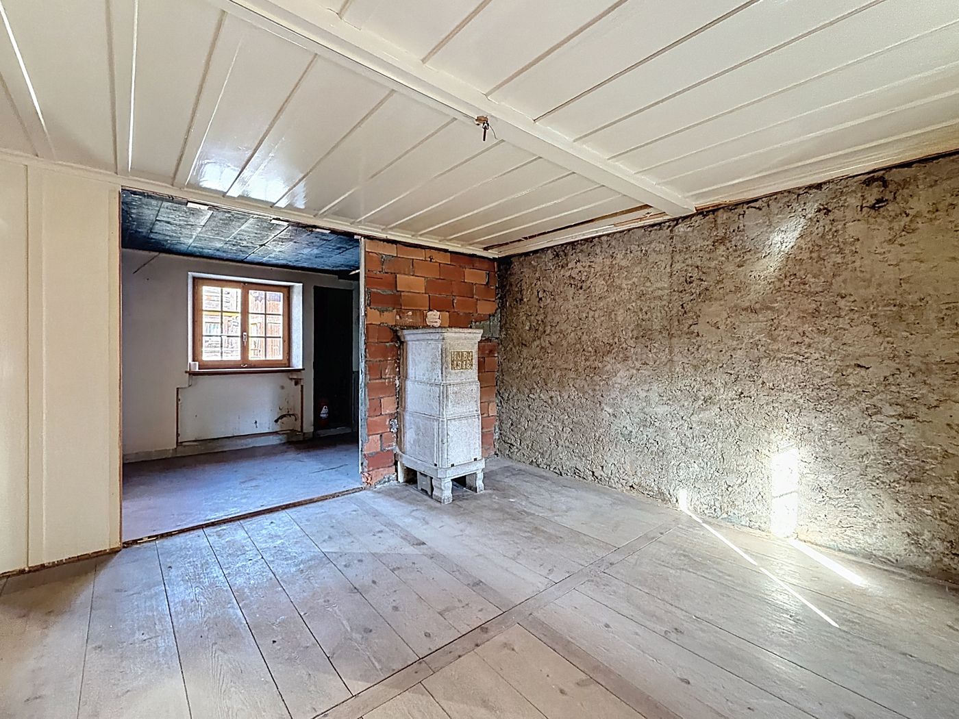 Pièce aux murs de pierre et plafond lambrissé blanc, poêle en faïence blanche, parquet brut, ouverture vers une seconde pièce.