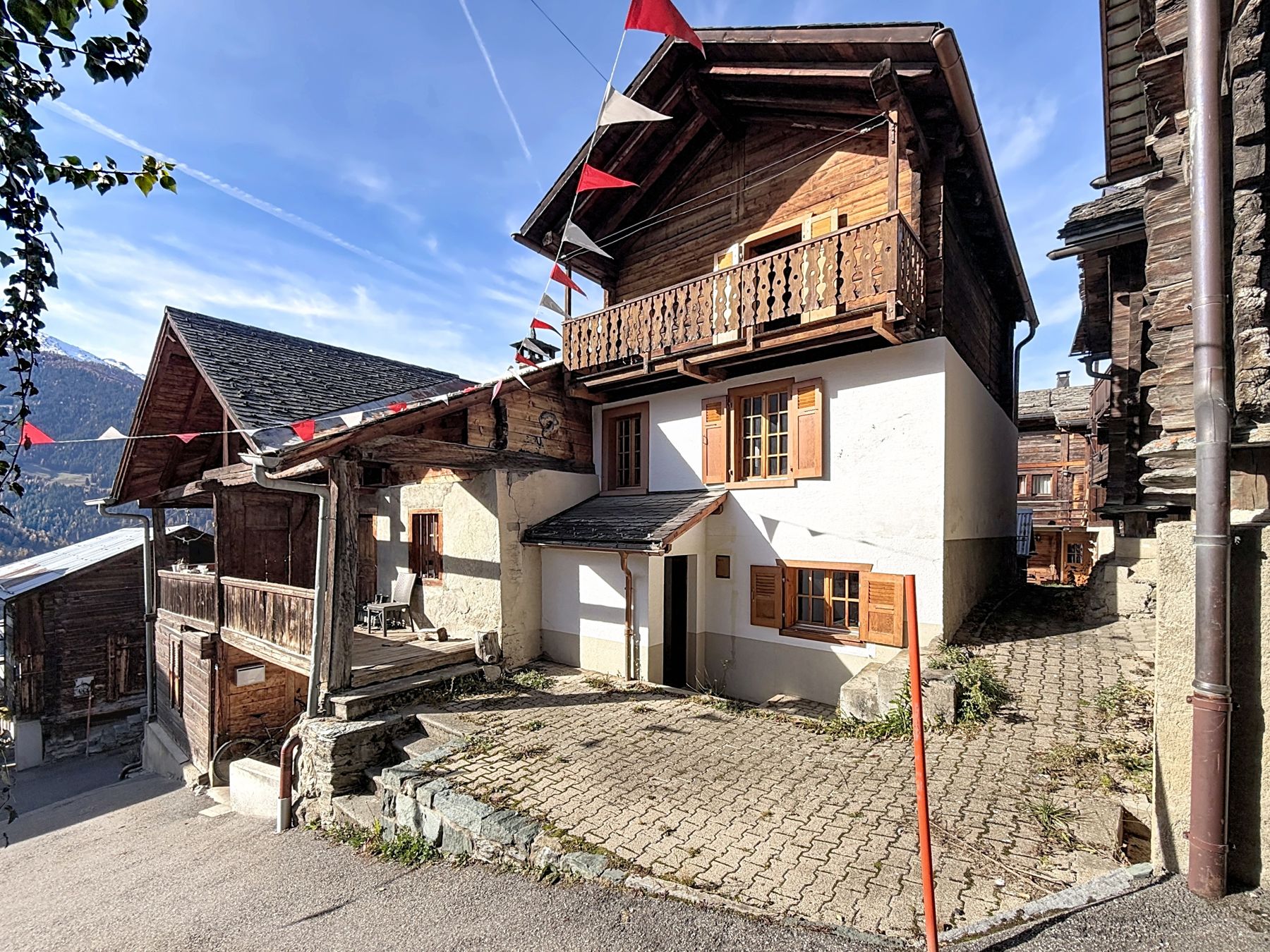 Le chalet Lucy au cœur de Sarreyer : maison crépie blanche aux volets de bois, étage en madriers bruns et balcon ouvragé, cour pavée, guirlande de fanions et ciel bleu.