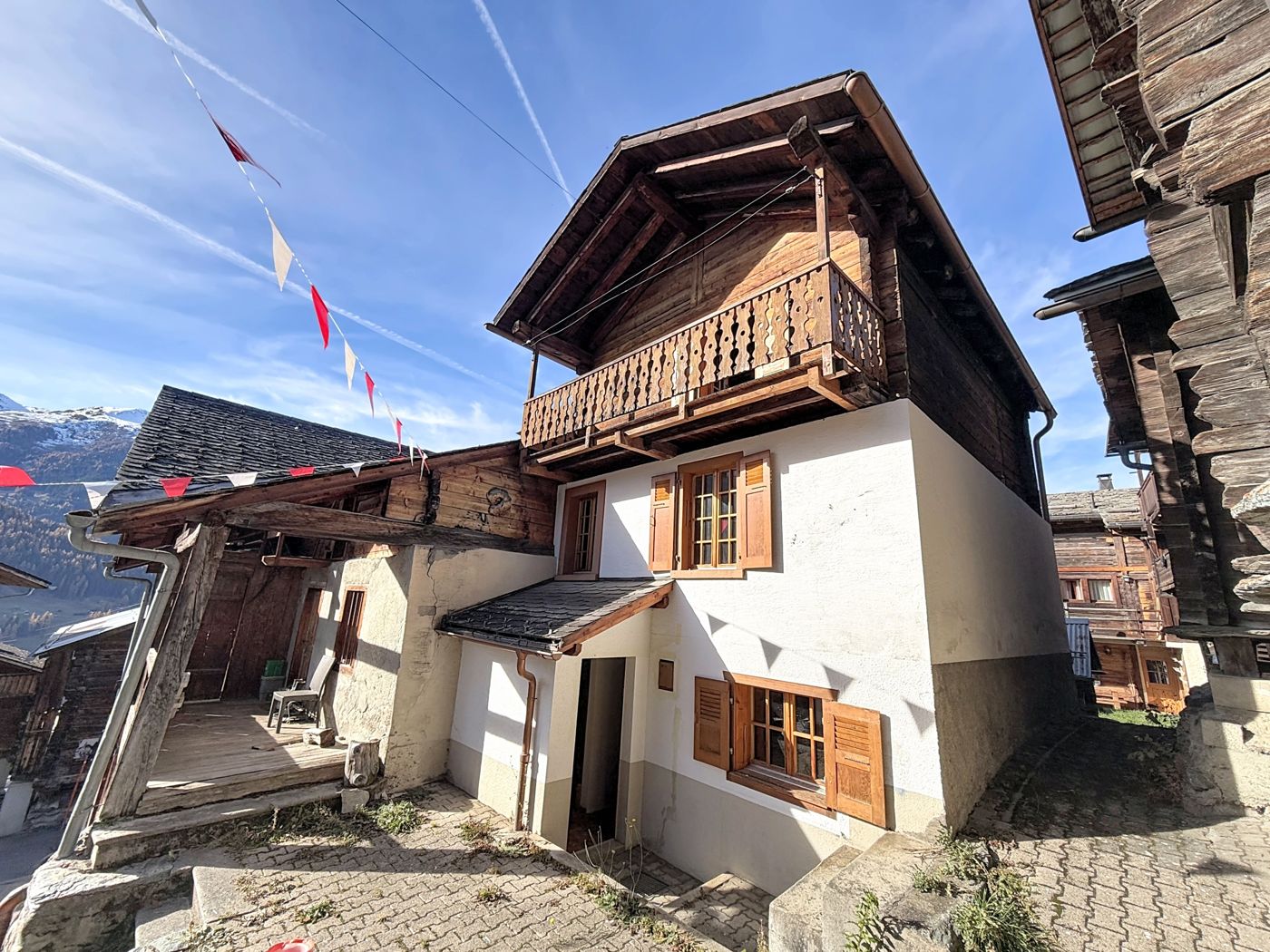 La maison vue de la ruelle : socle crépi blanc, étage en madriers et balcon, raccard voisin et vallée en arrière-plan.